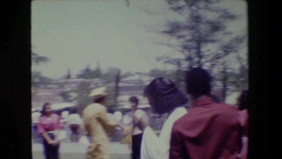 1983 Lansing home-movie frame showing several adults gathered outdoors near trees and a low stone wall in bright sun.