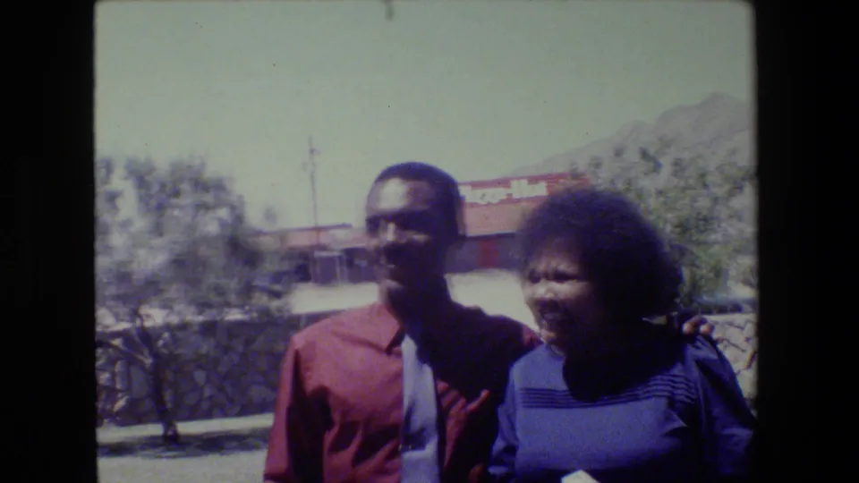 1983 Lansing frame with a man in a red shirt and light tie standing beside a woman in a blue striped top near a stone wall.