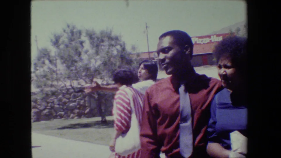 A man in a red shirt and tie and a woman walk past while another woman shields her face in the sunny 1983 Lansing scene.