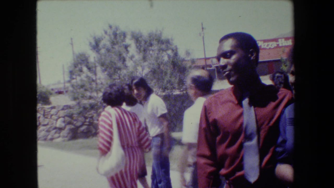 1983 Lansing home-movie frame showing African American adults walking outdoors near a visible Pizza Hut sign, with a smiling man in red closest to camera.