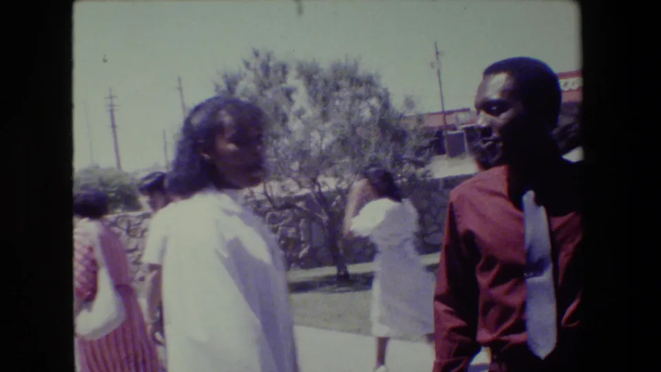 1983 Lansing frame with a woman in white moving past the camera and a man in a red shirt near the edge, with a Pizza Hut sign in the background.