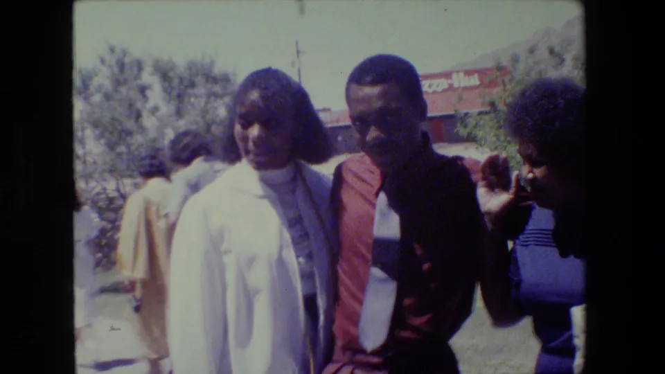 1983 Lansing home-movie frame showing adults clustered near the camera, including a woman in white, a man in a red shirt and tie, and a woman in blue.