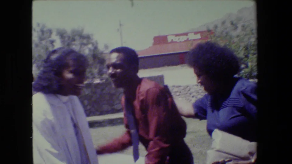 1983 Lansing frame showing a smiling man in a red shirt interacting with women in white and blue clothing outside near a Pizza Hut sign.