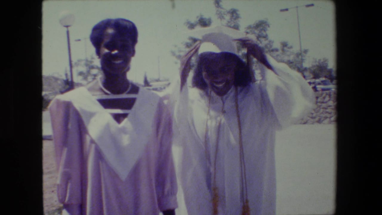Two young Black women in graduation caps and gowns smile outdoors in Lansing, Michigan, in 1983 home-movie footage.