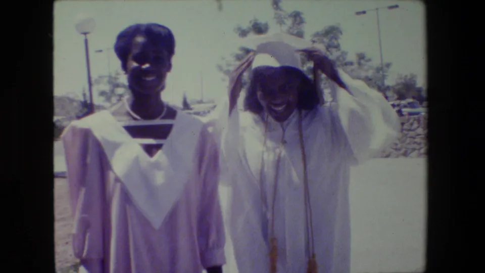 Two young Black women in graduation caps and gowns smile outdoors in Lansing, Michigan, in 1983 home-movie footage.