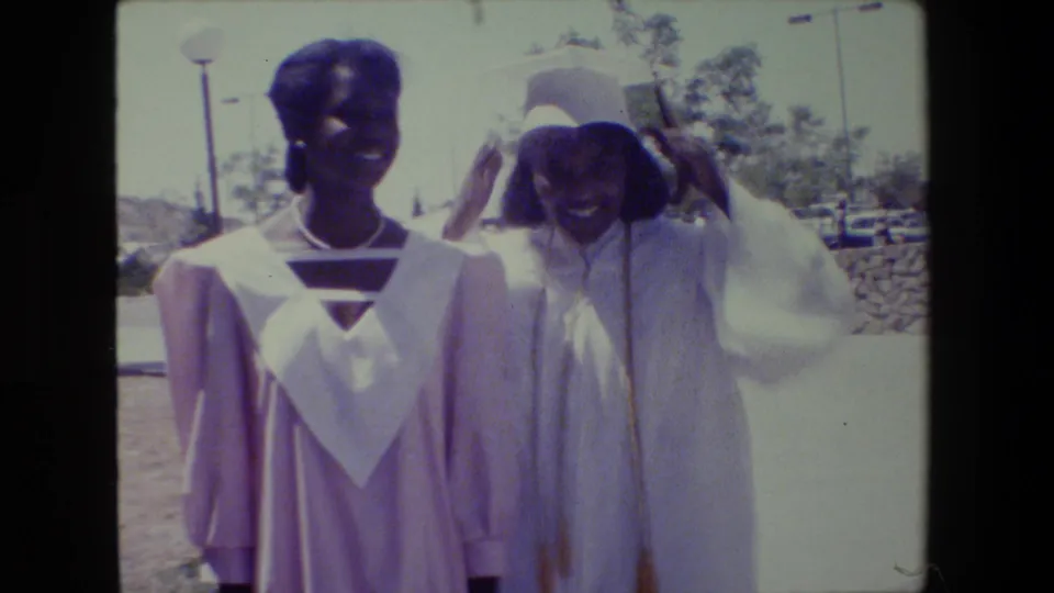 In the 1983 Lansing footage, two graduates stand side by side while the woman in white adjusts or lifts her cap.