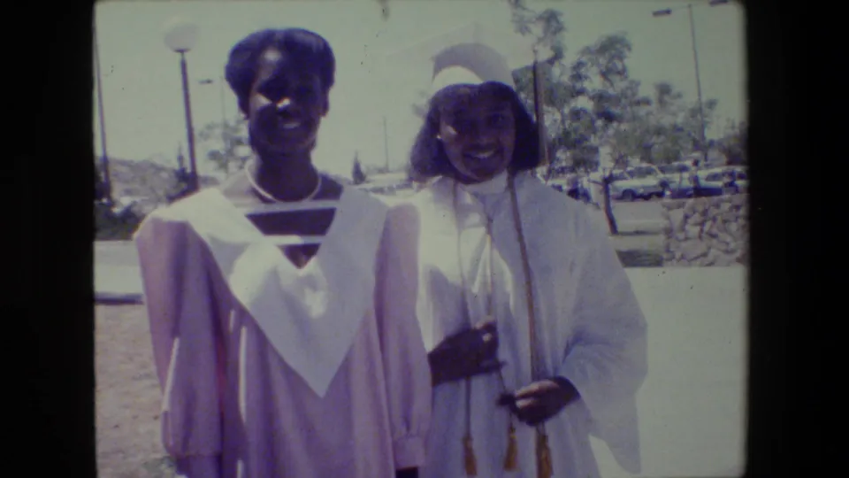 Two young Black women in graduation gowns smile together outdoors, with parked cars and trees behind them in Lansing, Michigan.