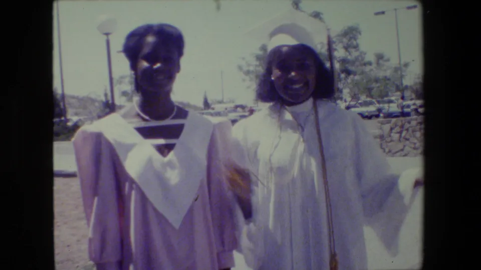 A smiling graduate in a white gown gestures beside another in lavender during the 1983 Lansing graduation celebration.