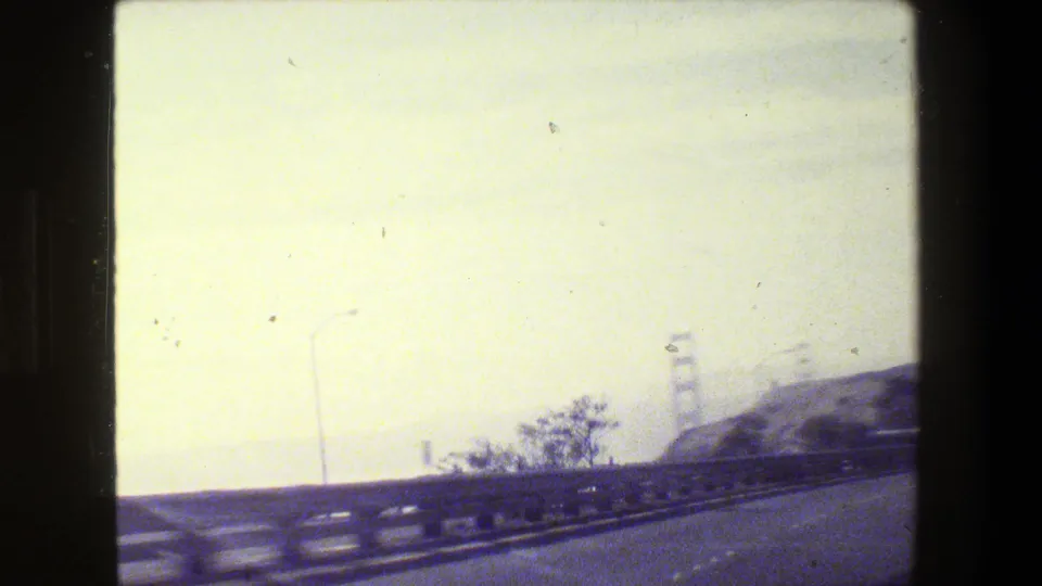 Moving-car view near San Francisco with freeway barrier, roadside trees, hills, and a hazy Golden Gate Bridge tower in the distance.