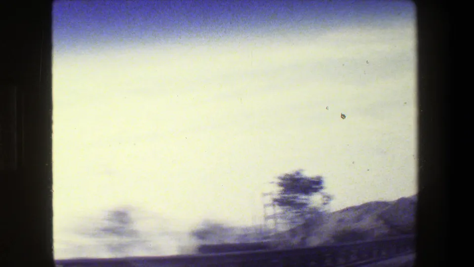 1984 8mm travel frame with motion-blurred roadside trees, freeway railing, hills, and a faint bridge tower near San Francisco.