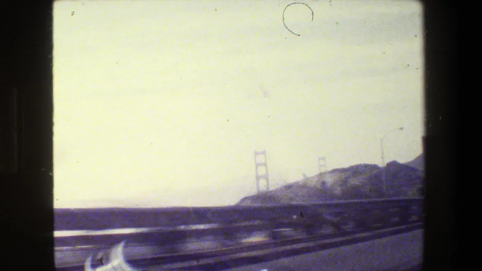 Hazy 1984 freeway approach toward the Golden Gate Bridge, with towers, hillside, guardrail, water, and 8mm dust marks visible.