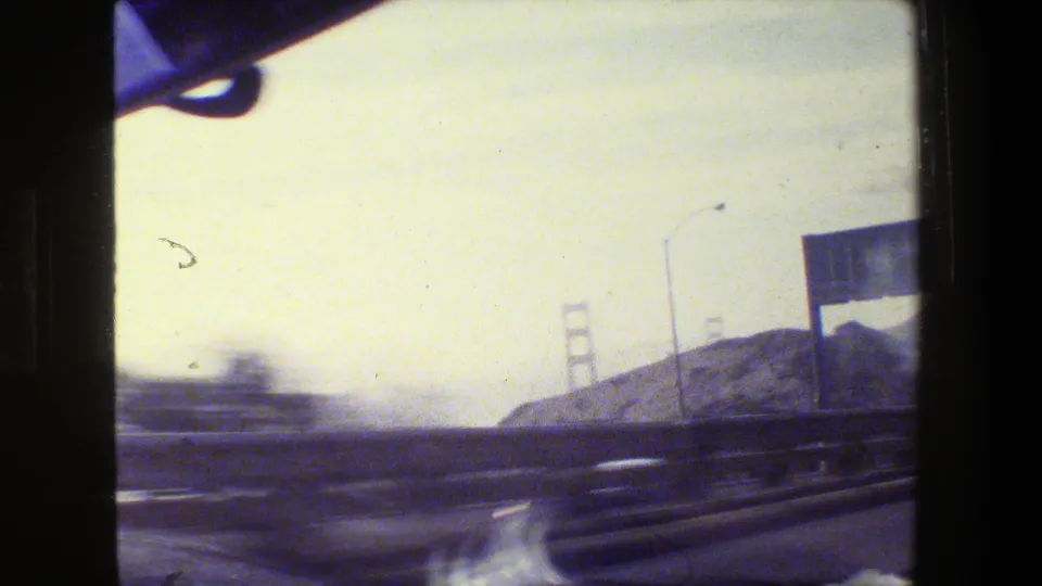 1984 San Francisco-area 8mm frame looking through a car windshield toward highway lanes, hills, and distant Golden Gate Bridge towers.