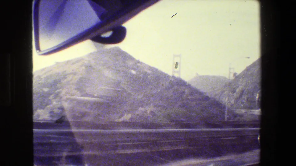 Moving 8mm car-window view in San Francisco showing a hillside, road barrier, rearview mirror, and Golden Gate Bridge towers in haze.
