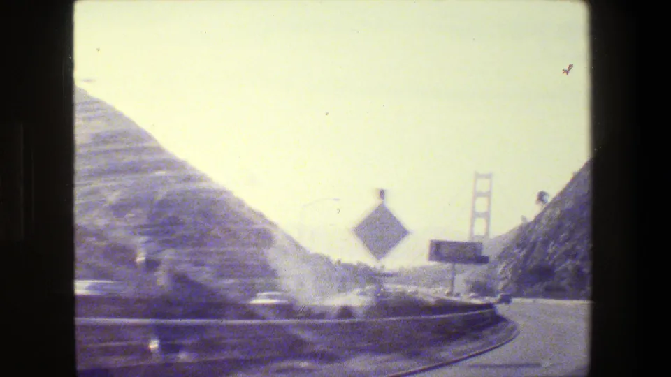 Final 1984 San Francisco-area frame showing roadside signs, sloping hills, blurred traffic, and a Golden Gate Bridge tower in the distance.