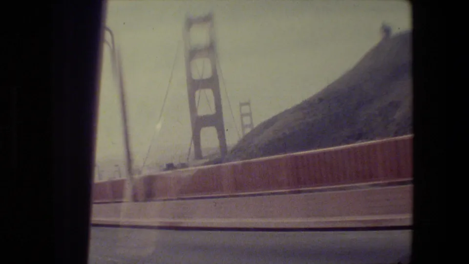 1984 home-movie frame from the Golden Gate Bridge approach, with towers, suspension cables, and red railing passing by.