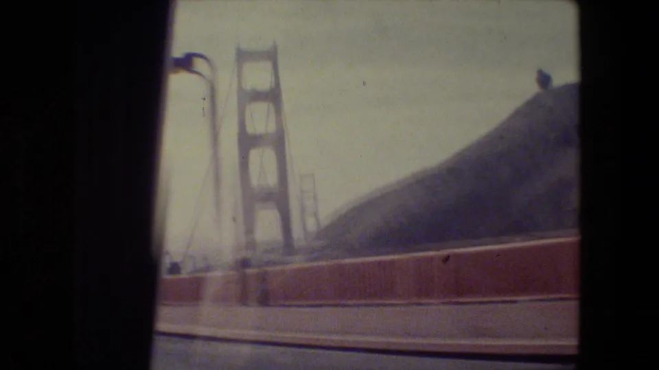 Close 1984 San Francisco bridge approach view, with Golden Gate Bridge tower, cables, railing, and hillside visible through motion blur.