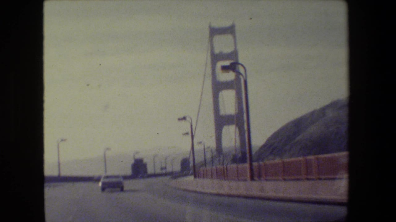 1984 8mm view from a car nearing a Golden Gate Bridge tower in San Francisco, with a car ahead, lamps, cables, and hillside visible.