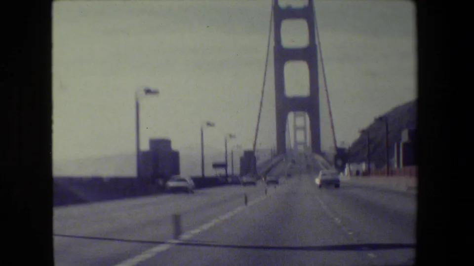 1980s 8mm roadway view from a moving car on the Golden Gate Bridge, with lanes, nearby cars, tower structure, lamps, and hillside.