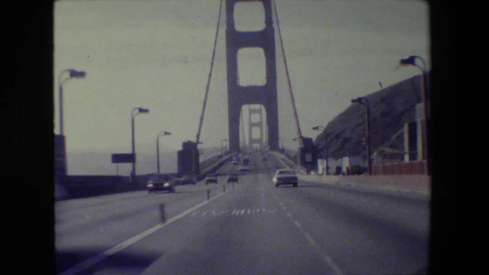 Forward-facing 1984 car view on the Golden Gate Bridge, showing traffic lanes, cars, red-orange railings, cables, lamps, and hills.