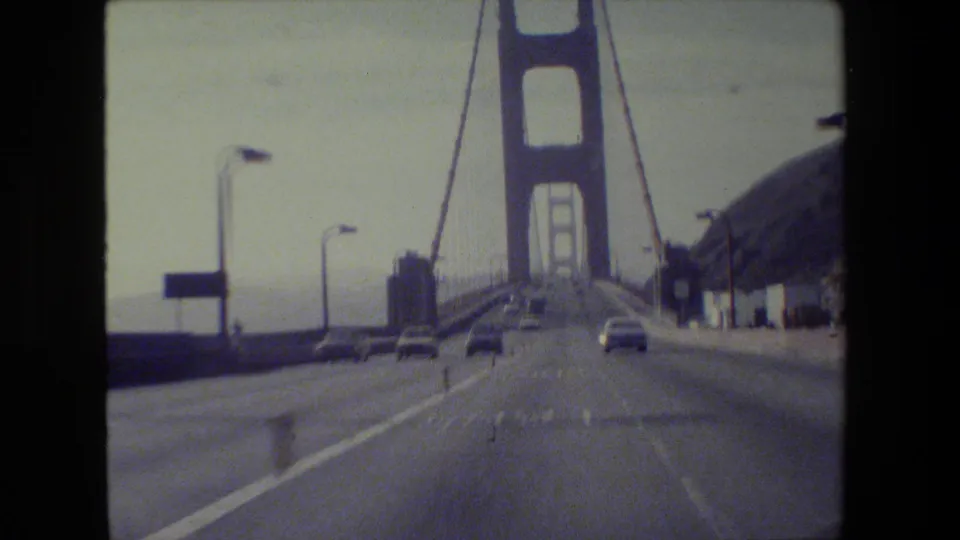 1984 San Francisco home-movie view from a vehicle approaching the Golden Gate Bridge, with cars, lanes, tower, and cables visible.