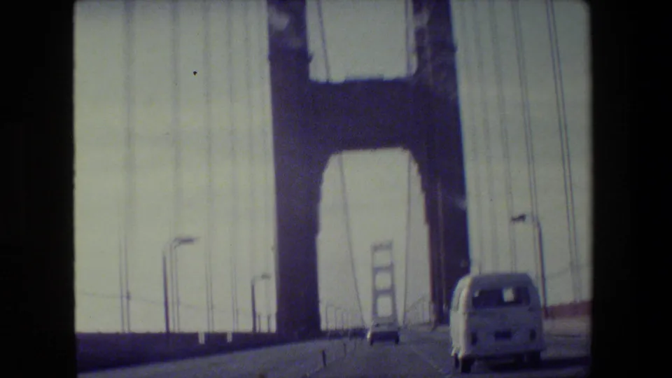 Driver’s-eye 1984 view on the Golden Gate Bridge, showing suspension cables, tower structure, cars, and a white van nearby.