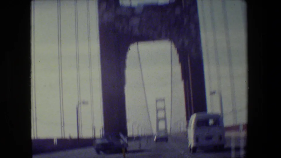 1984 home-movie driving shot on the Golden Gate Bridge, with the tower looming above traffic and suspension lines filling the frame.