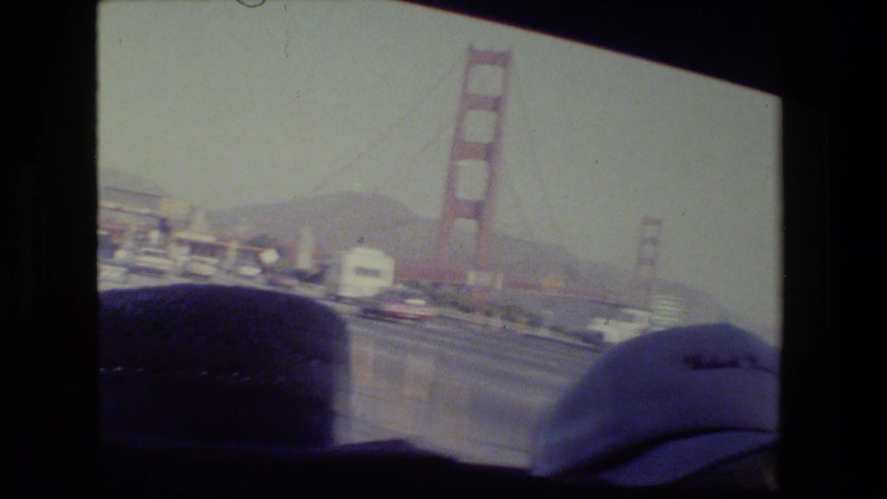 1984 San Francisco home-movie frame showing the Golden Gate Bridge from inside a moving car, with traffic and a passenger cap visible.