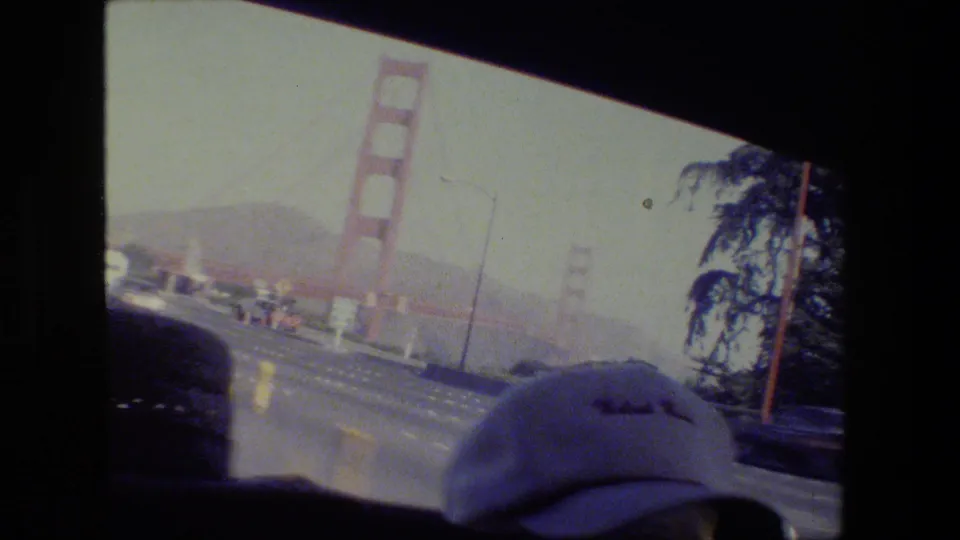 1984 San Francisco driving view toward the Golden Gate Bridge, filmed from a vehicle with traffic, roadside trees, and a foreground cap.
