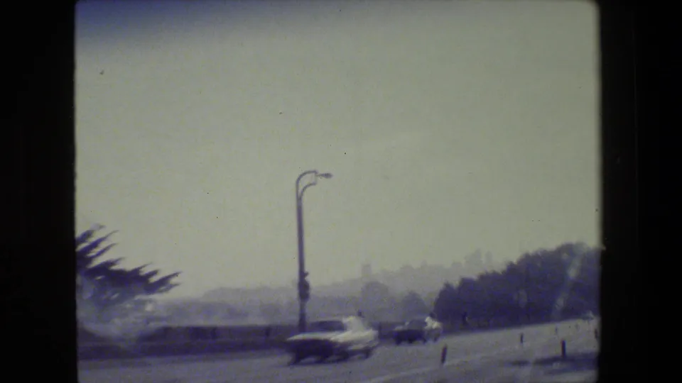 1984 San Francisco road scene from a moving vehicle, showing traffic, a tall street lamp, trees, and a hazy waterfront distance.