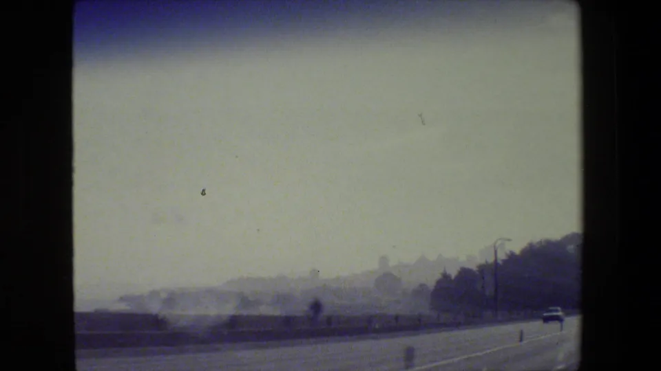 1984 San Francisco home-movie frame showing a hazy roadside waterfront view with trees, a lamp post, and sparse traffic.