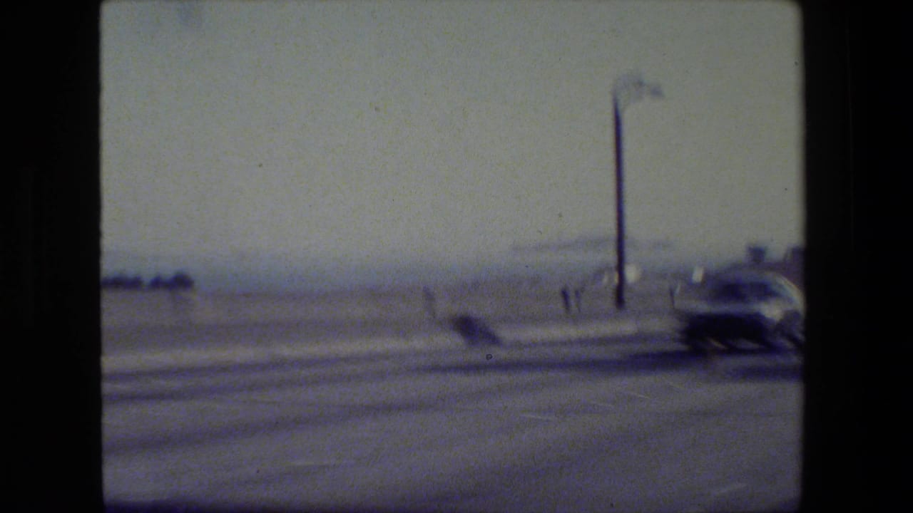1984 San Francisco 8mm frame from a moving vehicle showing a blurred road, a car, a streetlight, and hazy waterfront background.