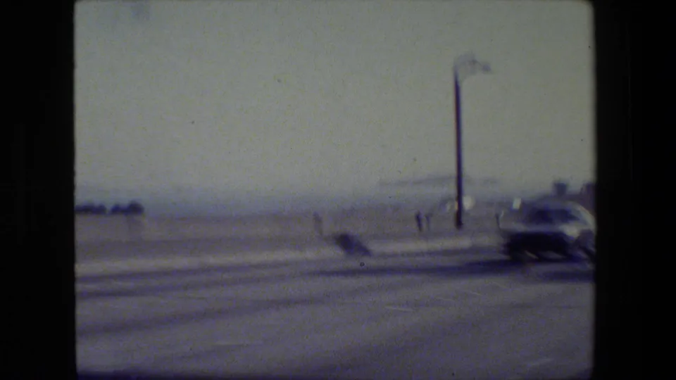 1984 San Francisco 8mm frame from a moving vehicle showing a blurred road, a car, a streetlight, and hazy waterfront background.