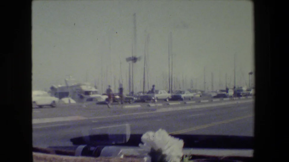 1984 San Francisco 8mm frame showing a road near a marina, with cars, sailboat masts, street fixtures, and the vehicle windshield edge.