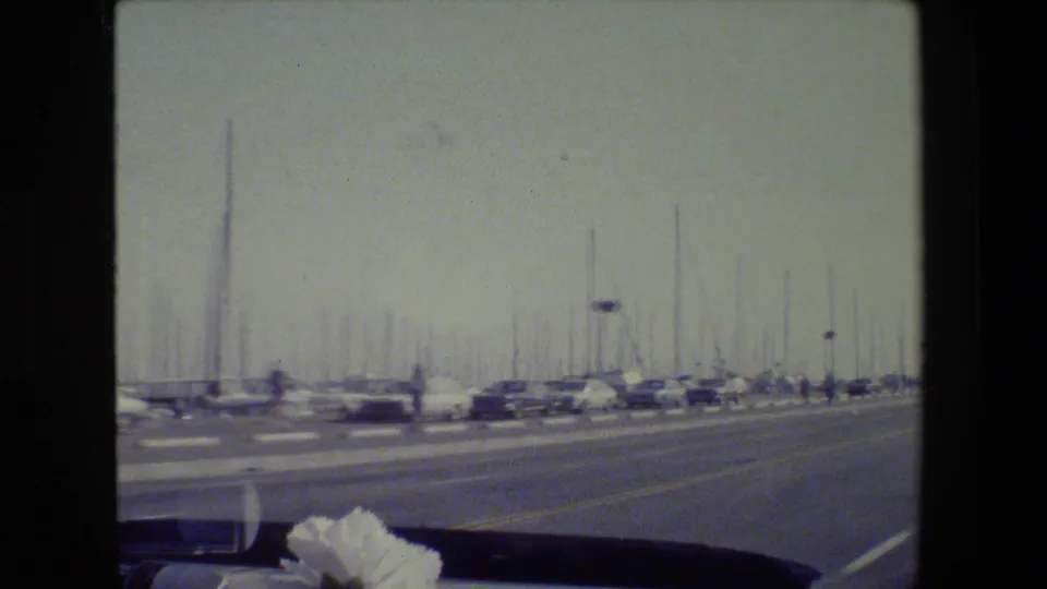1984 San Francisco 8mm frame looking past a roadway toward a marina with cars, dense sailboat masts, and a dark vehicle interior edge.