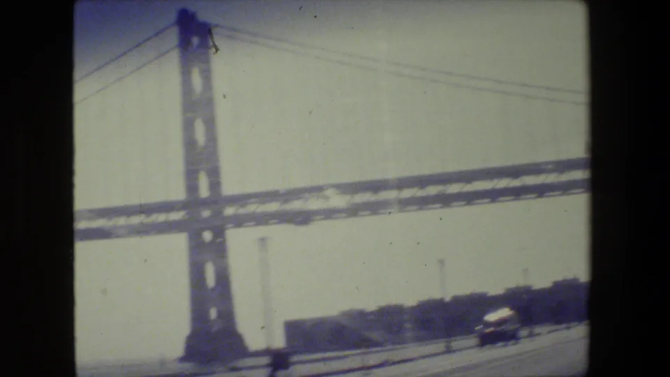 1984 San Francisco roadway view of a suspension bridge tower, cables, distant shoreline, and blurred traffic on 8mm film.