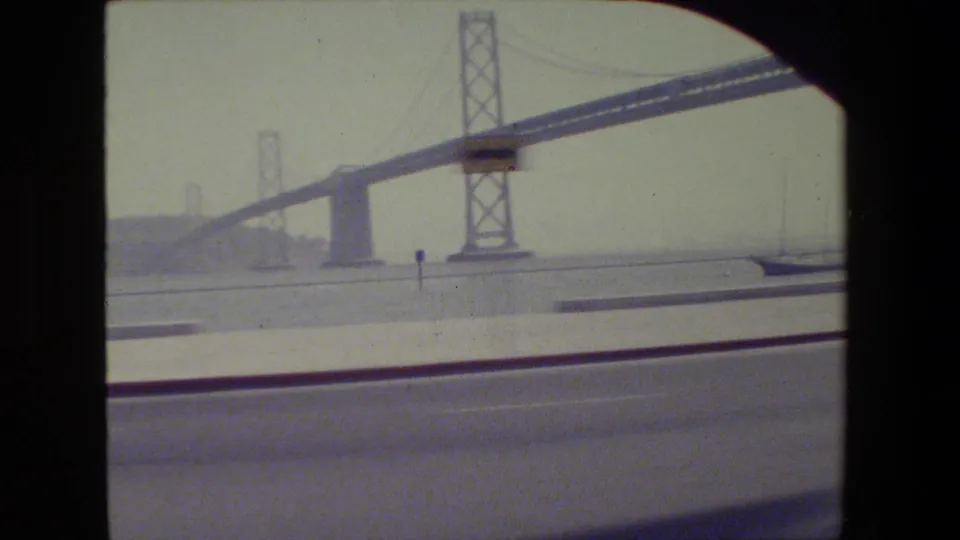 1984 San Francisco home-movie frame showing a large suspension bridge span, tower, roadway, and distant bay shoreline.