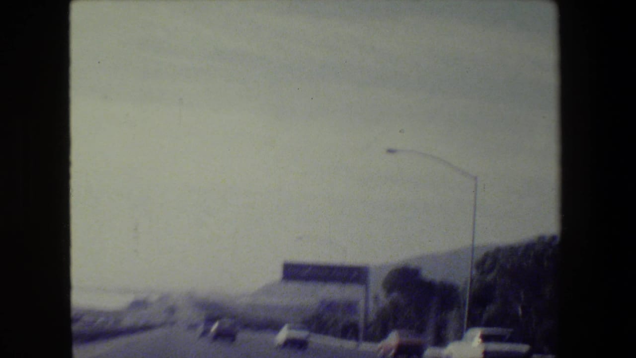 1984 San Francisco-area 8mm road-trip frame showing a freeway from a moving vehicle with traffic, hills, and a green guide sign.