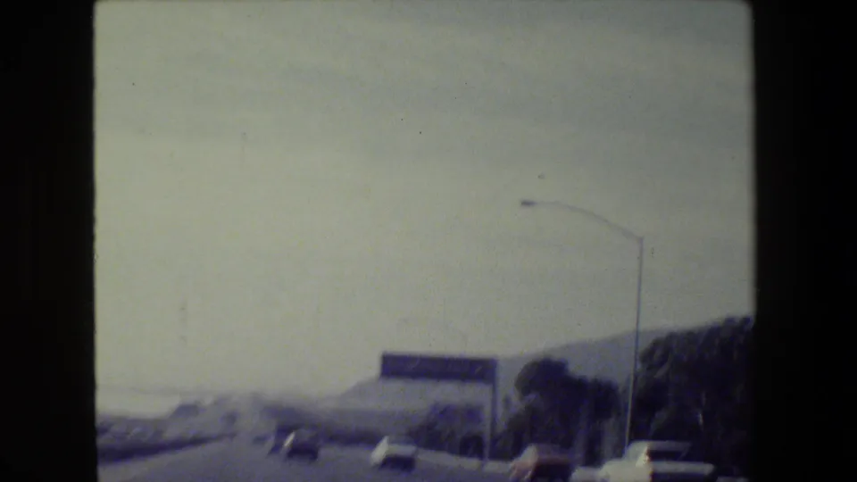 1984 San Francisco-area 8mm road-trip frame showing a freeway from a moving vehicle with traffic, hills, and a green guide sign.
