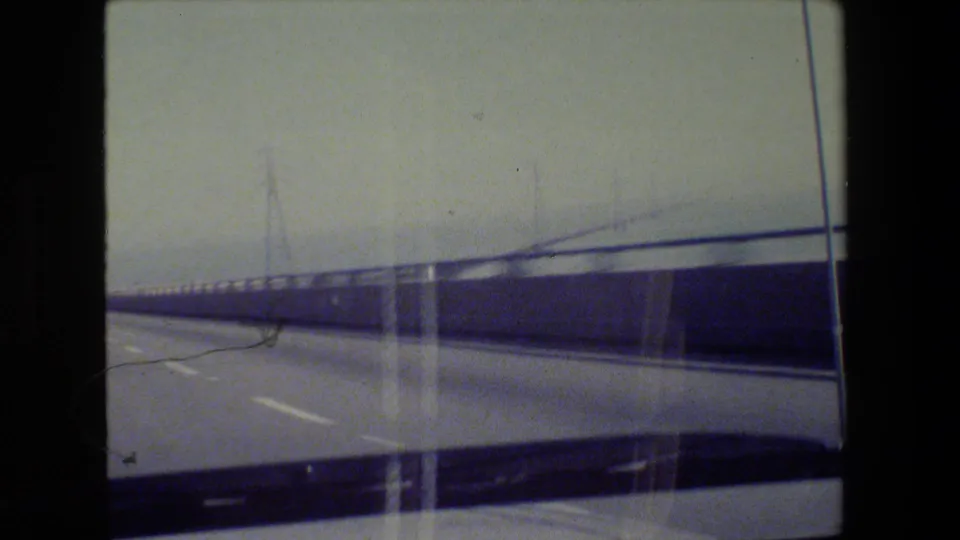 San Francisco-area 8mm frame from a moving vehicle crossing a long roadway or bridge with rails, poles, and hills beyond.