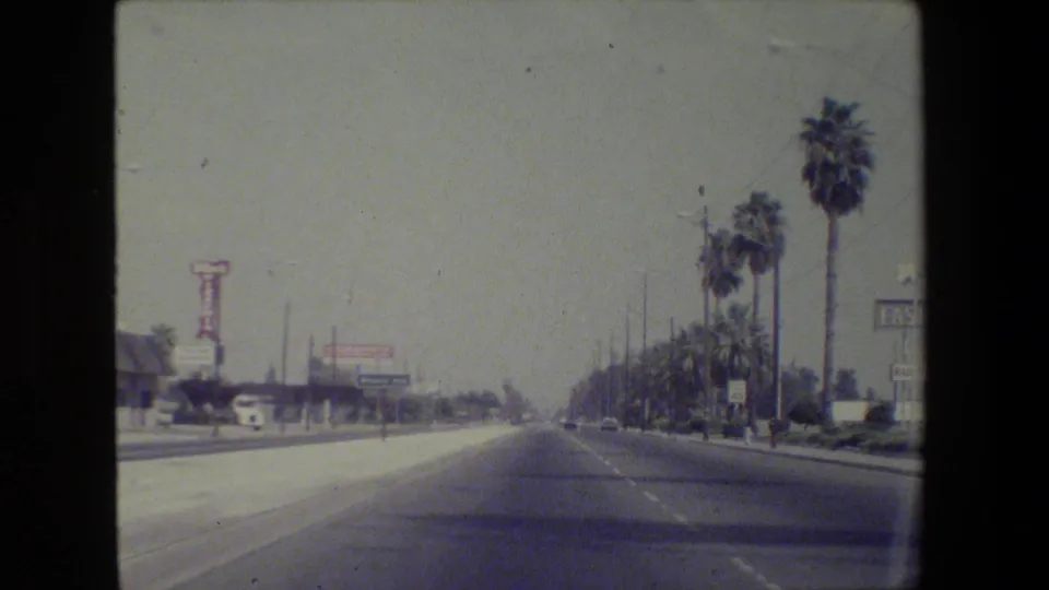 A 1984 8mm car-window view shows a multi-lane California road with palms, traffic lanes, and distant roadside signs.