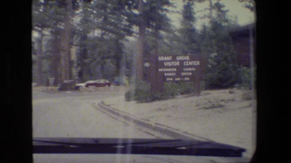 1980s 8mm frame showing a roadside Grant Grove Visitor Center sign, parked cars, trees, and the edge of a moving vehicle viewpoint.