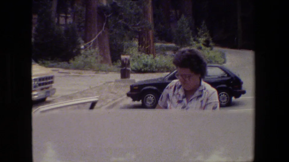 A woman with glasses stands near parked vehicles in a forest parking area in 1980s archival home-movie footage.