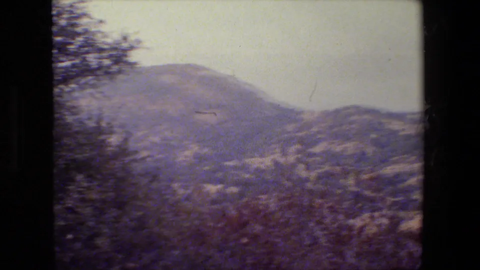 1984 home-movie frame showing rolling California hills and dry brush, with trees along the left side.