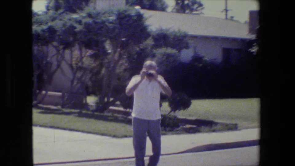 The older man appears to photograph the camera operator from the street, backed by shrubs, lawn, and a low house.