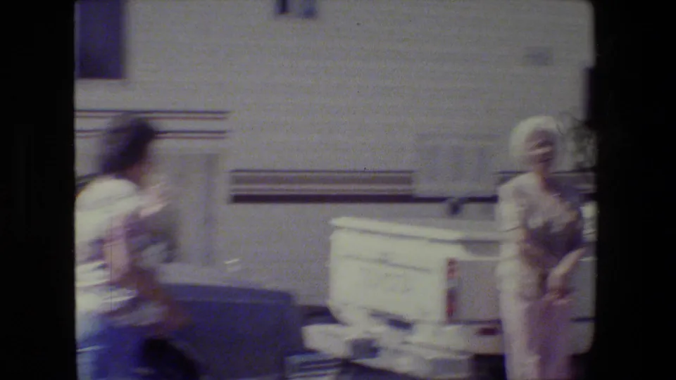 A person moves across the foreground as an older woman remains near the white pickup, with handheld blur and a pale building wall behind them.