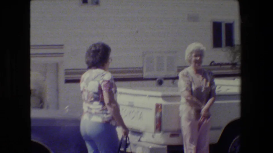 One woman in jeans and a patterned top faces an older woman near the white pickup, with the RV side and driveway visible in the background.