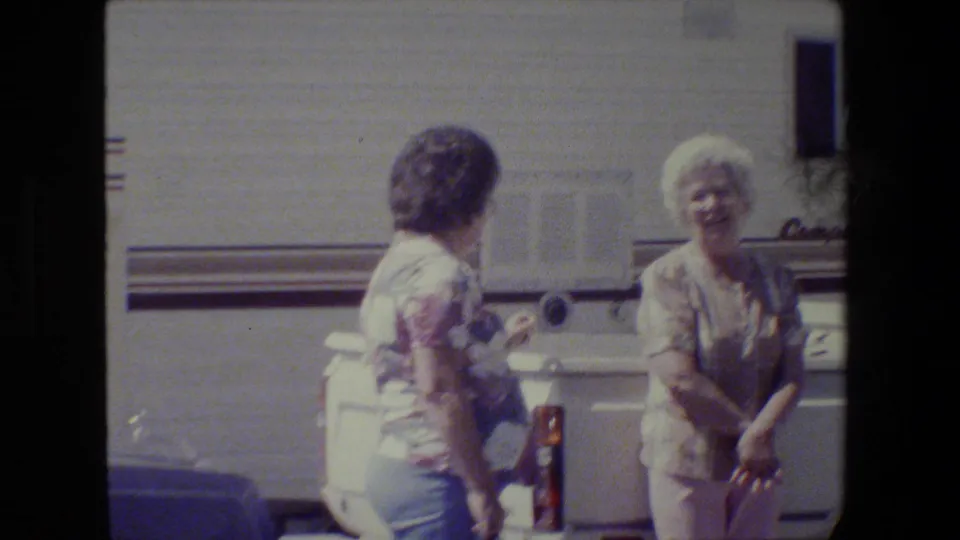 Two older women appear to converse beside the white pickup and RV, framed softly by faded 8mm home-movie color.
