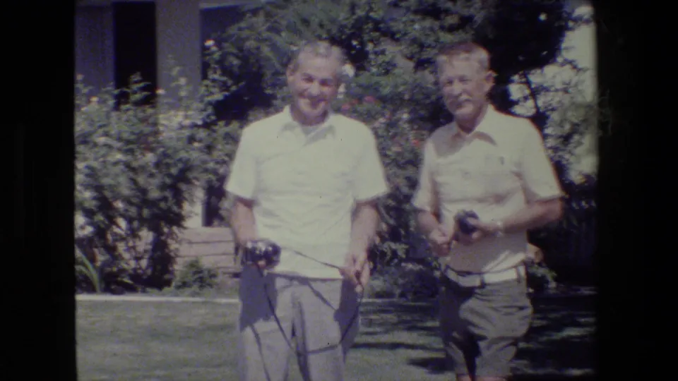 Two older men hold cameras while standing on a sunlit lawn near shrubs and a building, consistent with the 1984 San Francisco clip metadata.
