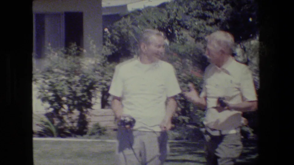 Two older men converse on a sunny lawn, with one holding a camera, in home-movie footage identified as San Francisco, California, 1984.