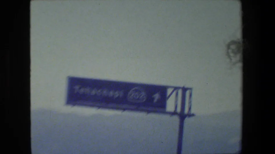 A soft, overexposed 8mm frame shows a partially readable exterior sign on a wall, with a cropped person at the right edge.